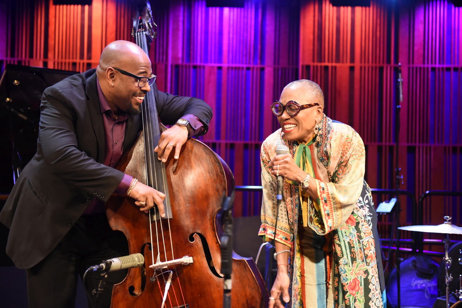 A smiling jazz vocalist and an upright bass player perform together on stage under warm purple and blue lighting. The singer holds a microphone and leans toward the bassist, both sharing a joyful moment mid-performance. A piano and drum set are visible in the background.
